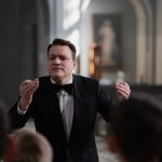 A male conductor leads a boys' choir rehearsal in a serene church setting.